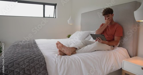 Man in early twenties tapping and swiping tablet on bed at home exploring content, copy space