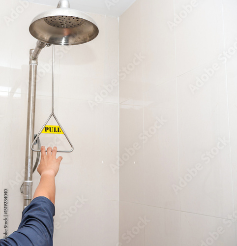 A person reaches to activate an emergency safety shower by pulling a triangular handle, demonstrating industrial safety protocols for chemical exposure or hazardous spills in the workplace.