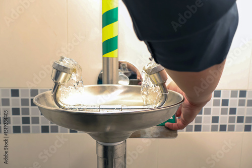 A person activates a stainless steel emergency eye wash station with dual nozzles in a tiled industrial setting, emphasizing workplace safety and hygiene precautions in hazardous environments.