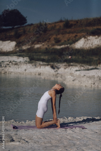Woman in white sportswear doing camel pose on yoga mat by lake. Outdoor flexibility and wellness practice. Design for poster, banner, greeting card, invitation. Side view