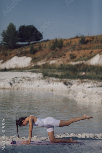 Woman in white sportswear practicing yoga on mat by lake in balancing position. Outdoor fitness and healthy lifestyle concept. Design for poster, banner, greeting card, invitation. Side view
