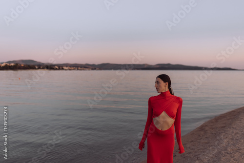 Woman in red cut-out dress standing on beach at sunset. Fashion photography. Summer vacation and travel concept. Design for poster, banner, greeting card, invitation. Side view with copy space