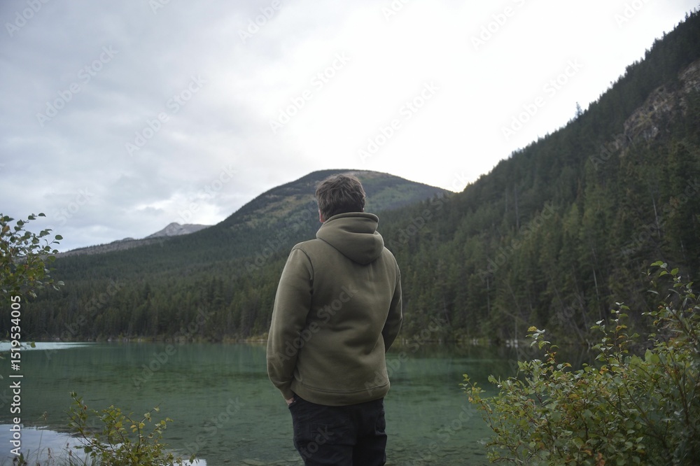 Fototapeta premium Man in a hoodie stands at the edge of a serene alpine lake.
