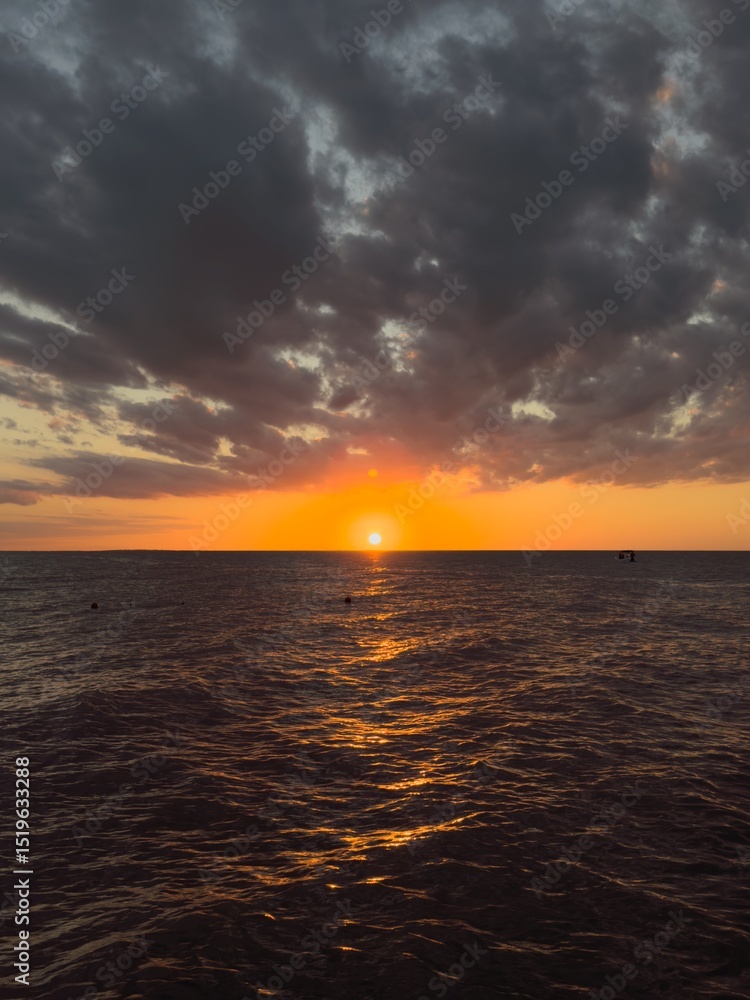 Fototapeta premium Vibrant red and orange sunset casting warm reflections on gentle ocean waves, photographed from the edge of a wooden pier. Serene and atmospheric seascape capturing the beauty of nature at dusk.