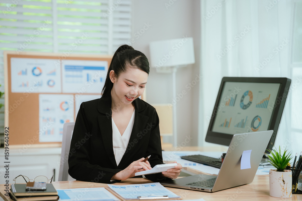 Confident Asian businesswoman smiling at her desk with laptop and financial charts, planning strategy and analyzing data for business success. Confident Asian businesswoman smiling at her desk with laptop and financial charts, planning strategy and analyzing data for business success.