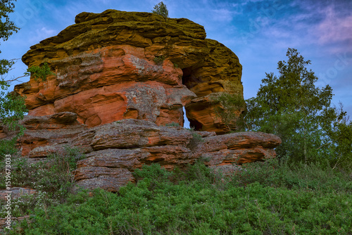 A huge stone rock with a hole in the green jungle with lush vegetation at dawn, flooded with the morning sun. Kazakh expanses, Mount Ku Egendybulak. Summer autumn landscape