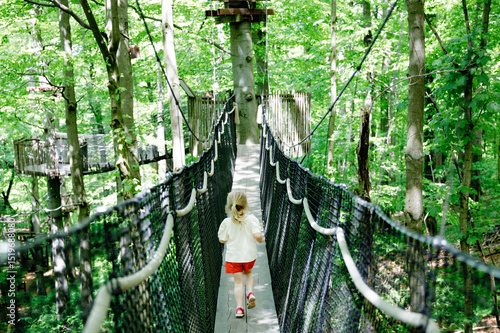 Photography Little preschool girl walking on high tree-canopy trail with wooden walkway and ropeways on Hoherodskopf in Germany