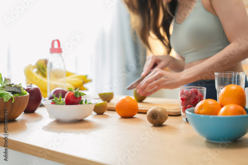 Healthy Asian woman in activewear prepares a fresh fruit drink, slicing fruit and blending a protein shake in her modern pantry kitchen at home, promoting wellness and fit lifestyle