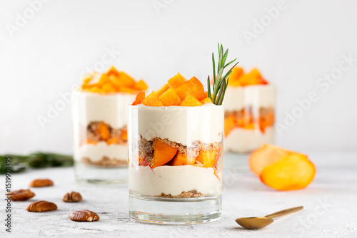 Parfait in a glasses with persimmon, pecan, whipped cream and biscuit on a light table. Healthy food, vegan, sugar, gluten and lactose free.