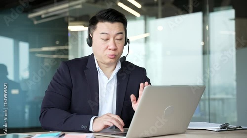 Asian male corporate operator in headset working in customer support service on helpline telesale talking using laptop in business office. Agent of call center advises client answering questions