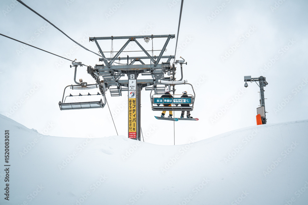 A snowboarders ride a ski lift over a snow-covered slope at Gala Yuzawa, Japan. The minimal backdrop and cold atmosphere highlight the ascent toward adventure on this peaceful winter day. A snowboarders ride a ski lift over a snow-covered slope at Gala Yuzawa, Japan. The minimal backdrop and cold atmosphere highlight the ascent toward adventure on this peaceful winter day.