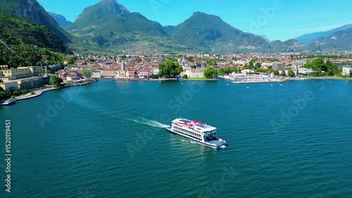 Aerial view of Riva del Garda with Torre Apponale, MAG museum, bike paths, boats and river ferries connecting towns, surrounded by mountains and clear Lake Garda water, full of tourists