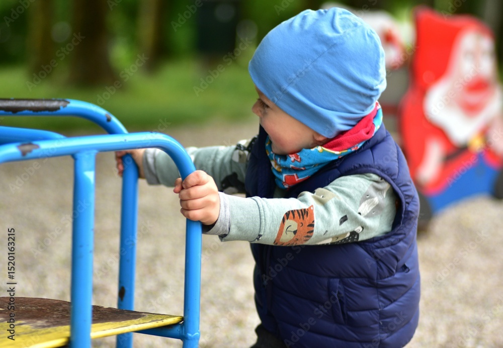Obraz premium Smiling toddler boy playing at a playground, holding on to colorful bars. Dressed warmly in a beanie and vest, he enjoys outdoor playtime on a chilly day surrounded by nature.