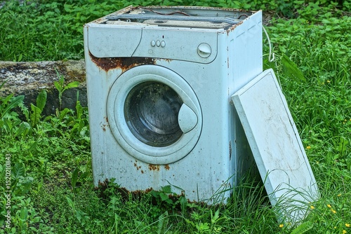 one industrial closed rusty dirty iron white broken old square modern electric with cover front discarded washing machine standing on green grass daytime	