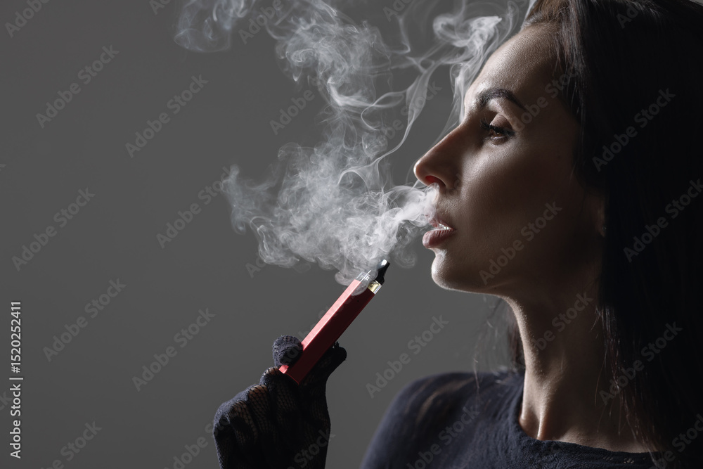 Obraz premium Side profile of a woman vaping with a sleek red electronic cigarette, exhaling thick vapor in a moody setting. Stylish and modern concept of vaping lifestyle and addiction.