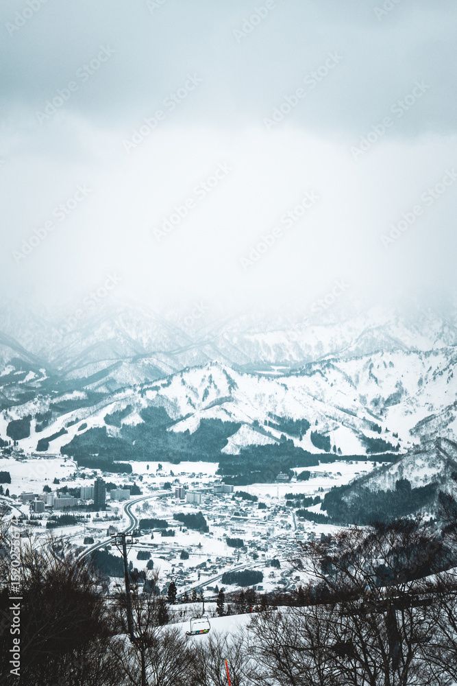 Obraz premium Skiers enjoy the slopes of Gala Yuzawa with stunning views of Ishiuchi Maruyama and Yuzawa Kogen in Niigata, Japan. Snow-covered mountains and a small town stretch across a scenic winter valley.