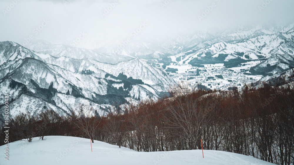 Obraz premium Skiers enjoy the slopes of Gala Yuzawa with stunning views of Ishiuchi Maruyama and Yuzawa Kogen in Niigata, Japan. Snow-covered mountains and a small town stretch across a scenic winter valley.