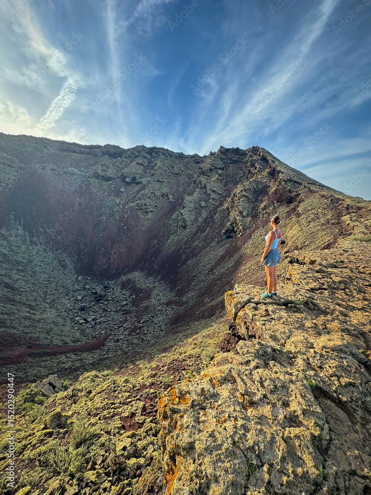 Fototapeta premium Woman stands on the rugged edge of a vast volcanic crater, gazing into its depths under a partly cloudy sky. Dramatic landscape shows immense scale and raw power of the natural geological formation.