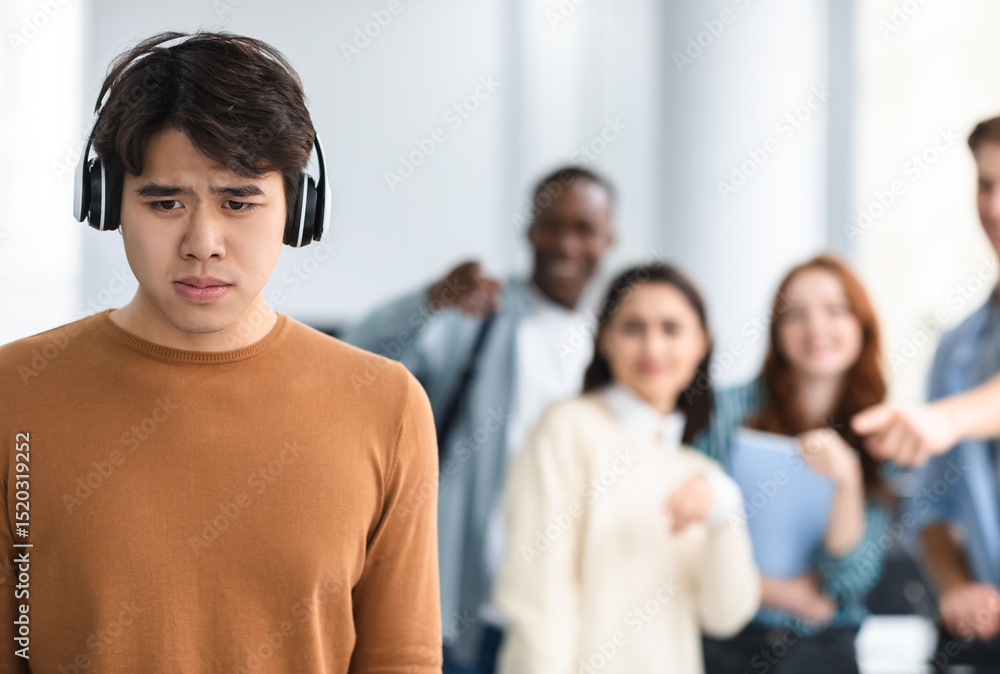 Obraz premium Group Conflict. Sad asian guy in headphones standing away alone, suffering humiliation and public disgrace, crowd of bad people bullying pointing fingers at victim, laughing. Racial Discrimination