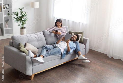 High angle view of relaxed cheerful stylish millennial eastern lovers man and woman chilling on couch in cozy living room, using gadgets at home, scrolling on social media, reading online, copy space