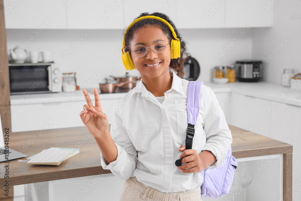 Naklejka premium Little African-American schoolgirl with headphones and backpack showing victory gesture in kitchen