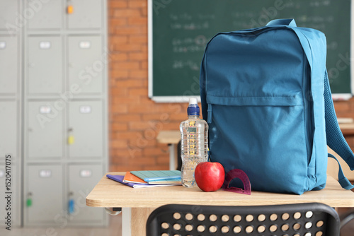 Blue backpack with apple, water bottle and copybooks on desk in classroom