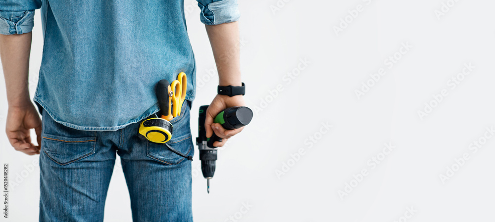 Men work, home renovation, construction or carpentry business. Young guy in jeans holds electric drill, with tools in his pocket, isolated on white background, panorama, empty space, cropped, back Men work, home renovation, construction or carpentry business. Young guy in jeans holds electric drill, with tools in his pocket, isolated on white background, panorama, empty space, cropped, back