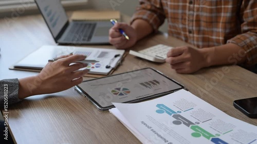 Two people are sitting at a table with a laptop, a calculator, and a stack of papers. They are discussing something important, possibly related to business or finance. The atmosphere seems serious