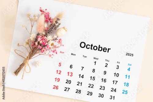 Close-up of paper sheet of calendar for october 2025 with beautiful small bouquet of dried flowers on beige table.