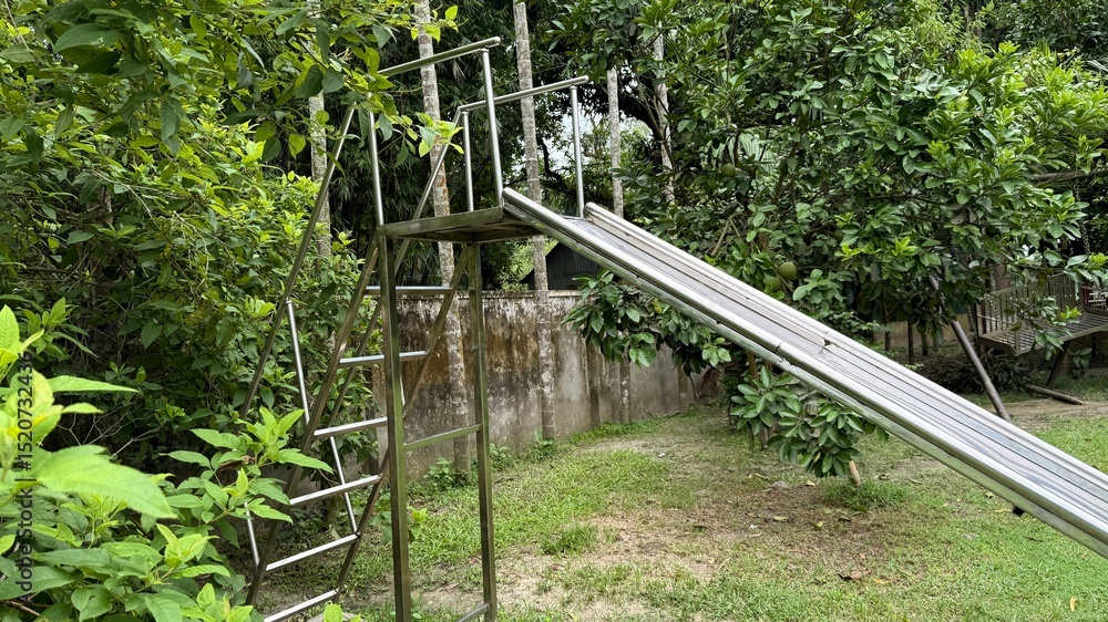 Silver Metal Slide with Ladder in Overgrown Backyard Surrounded by Lush Greenery Silver Metal Slide with Ladder in Overgrown Backyard Surrounded by Lush Greenery