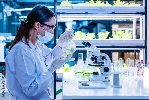 Female scientist examines green solution in a lab surrounded by plants and test tubes, representing eco-friendly farming innovations, sustainable agriculture, and plant-based biotechnology research.