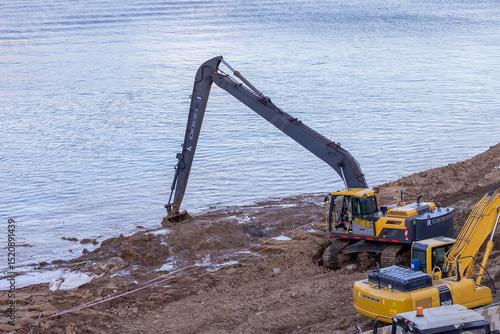 Long-arm excavator dredging riverbank with construction equipment nearby.