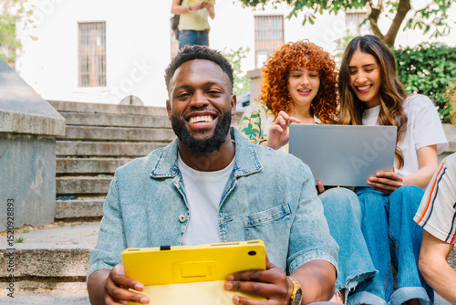 Diverse university students sitting on campus stairs, working on digital tablets and laptops, engaging in collaborative learning through technology