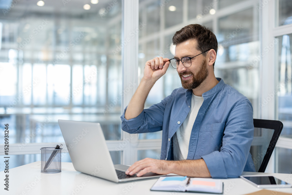 A smiling businessman in glasses works on a laptop in a bright office setting, possibly editing or working from home. A smiling businessman in glasses works on a laptop in a bright office setting, possibly editing or working from home.