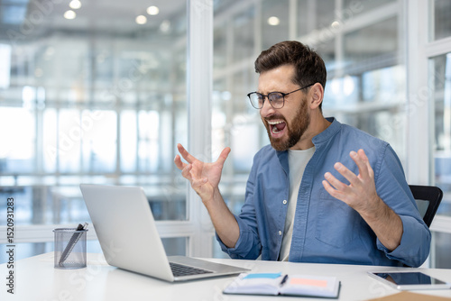 Papier peint An exasperated businessman, wearing glasses, shouts in frustration while working on his laptop at his office desk