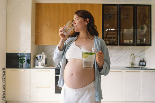 In a contemporary kitchen, a pregnant woman with a joyful expression holds a pitcher filled with water and a glass. Bright light enhances her radiant smile as she enjoys hydration.