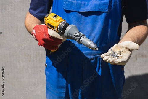 A man in a blue jumpsuit holds electric screwdriver with a riveter attachment and exhaust construction rivets on the background of a slate wall