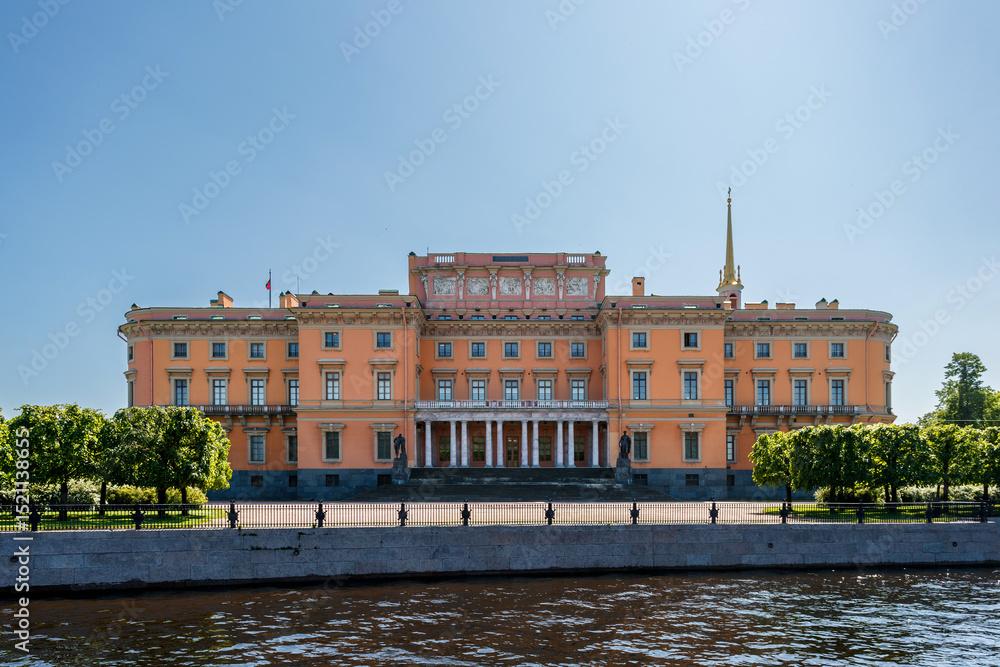 Fototapeta premium Mikhailovsky Castle of pink color with columns and a spire, stands on the river bank under a clear sky.