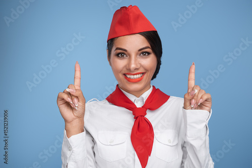 Fotografi Professional flight attendant in uniform smiling warmly, gesturing with raised hands against a solid blue background, symbolizing helpfulness, friendliness, and excellent customer service