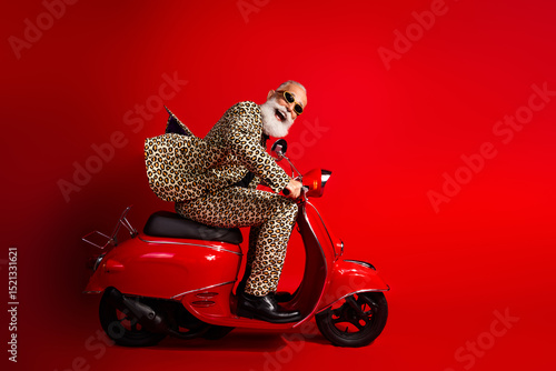 Fashionable elderly man wearing leopard print suit rides red scooter against red background exuding humor and eccentric style