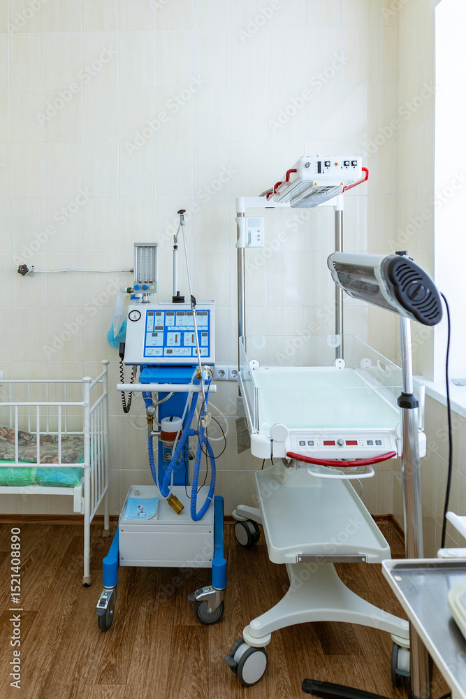 A clean, empty hospital incubator room features a white newborn baby crib next to an infant warmer and a ventilator. A clean, empty hospital incubator room features a white newborn baby crib next to an infant warmer and a ventilator.
