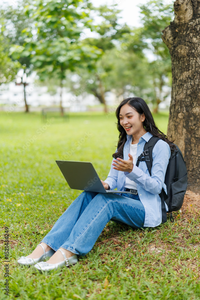 Happy Asian student sitting under a tree in a park, using her laptop for a video call, smiling and gesturing joyfully while enjoying the beauty of nature and the outdoors Happy Asian student sitting under a tree in a park, using her laptop for a video call, smiling and gesturing joyfully while enjoying the beauty of nature and the outdoors