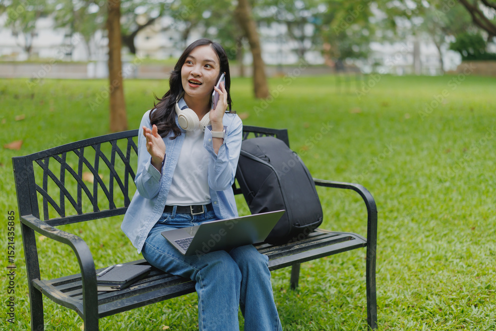 Asian freelancer woman is working on her laptop and talking on phone, sitting on bench in park, enjoying good weather and fresh air while working outdoors Asian freelancer woman is working on her laptop and talking on phone, sitting on bench in park, enjoying good weather and fresh air while working outdoors
