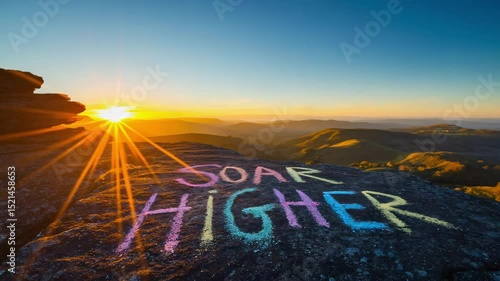 A colorful message 'Soar Higher' written in chalk on a rocky surface during sunset. The sky is filled with vibrant hues and distant hills are visible.