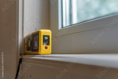 Yellow laser distance measurer placed on a window sill, its digital screen illuminated, ready to measure dimensions for installation or inspection.