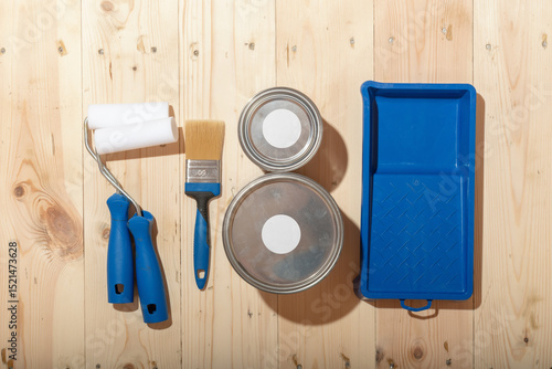 Fototapeta Set of paint rollers, brush, paint cans and blue tray neatly arranged on clean wooden floor, suggesting careful preparation for renovation