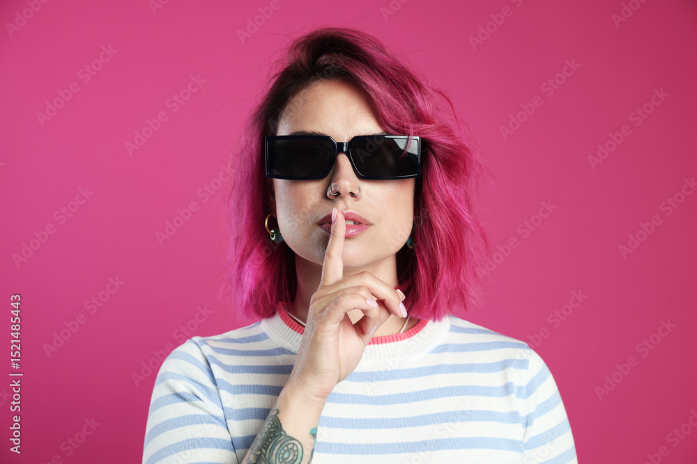 Fototapeta premium Beautiful woman with bright dyed hair in sunglasses making hush gesture on pink background