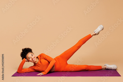 Canvas Print Full body young fitness trainer instructor sporty woman wear orange clothes spend time in gym do side leg raise exercise on yoga mat isolated on plain beige background