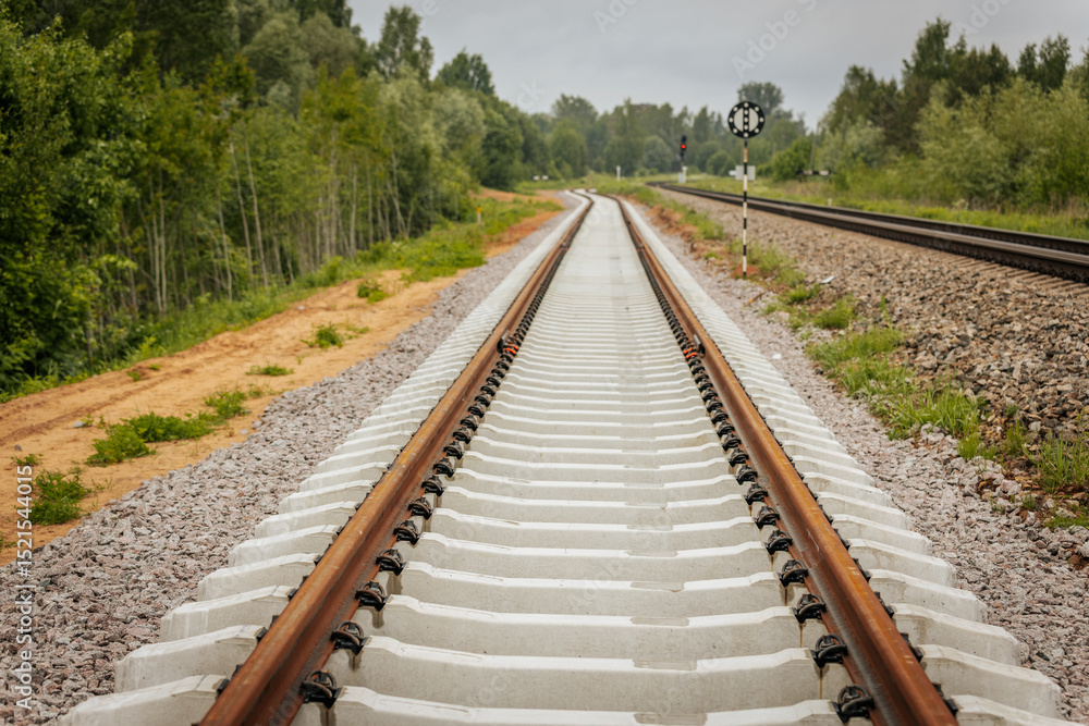 Obraz premium A curved section of newly laid railway track diverges beside an older line, with a circular signal sign and red light signal post in a green, forested area.