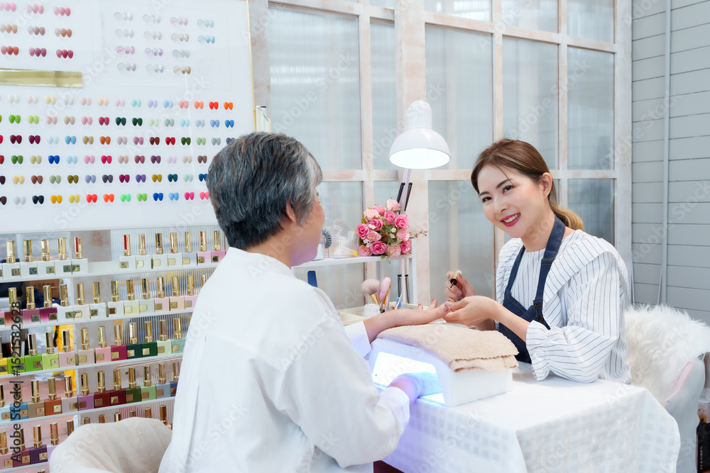 Obraz premium Senior Asian woman receiving manicure service from young Asian female nail technician smiling warmly in cozy small nail spa shop decorated with flowers and nail polish sample display